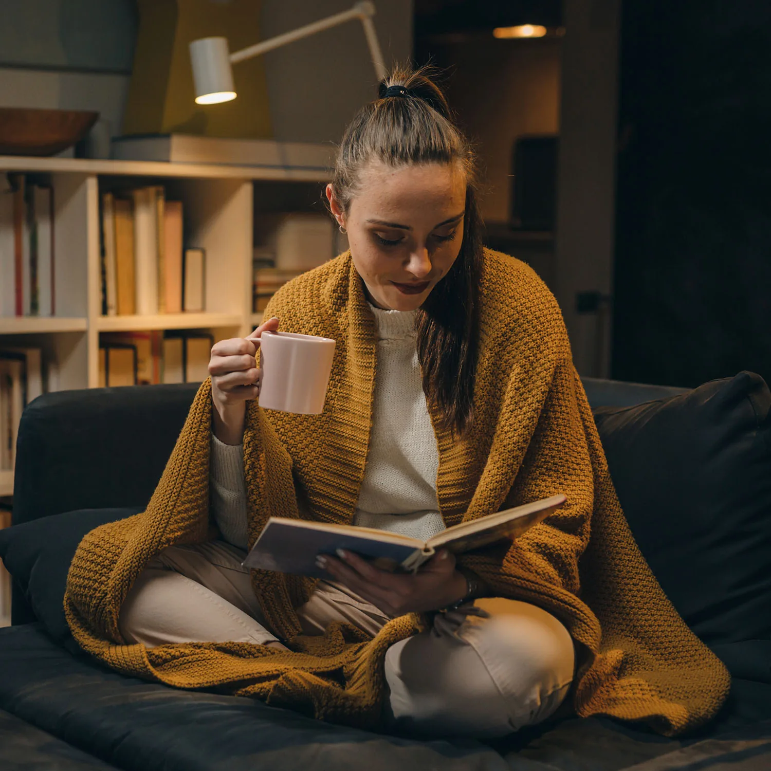 Eine Frau sitzt auf dem Sofa, eingekuschelt in eine Strickdecke, liest ein Buch und hält eine Tasse in der Hand.
